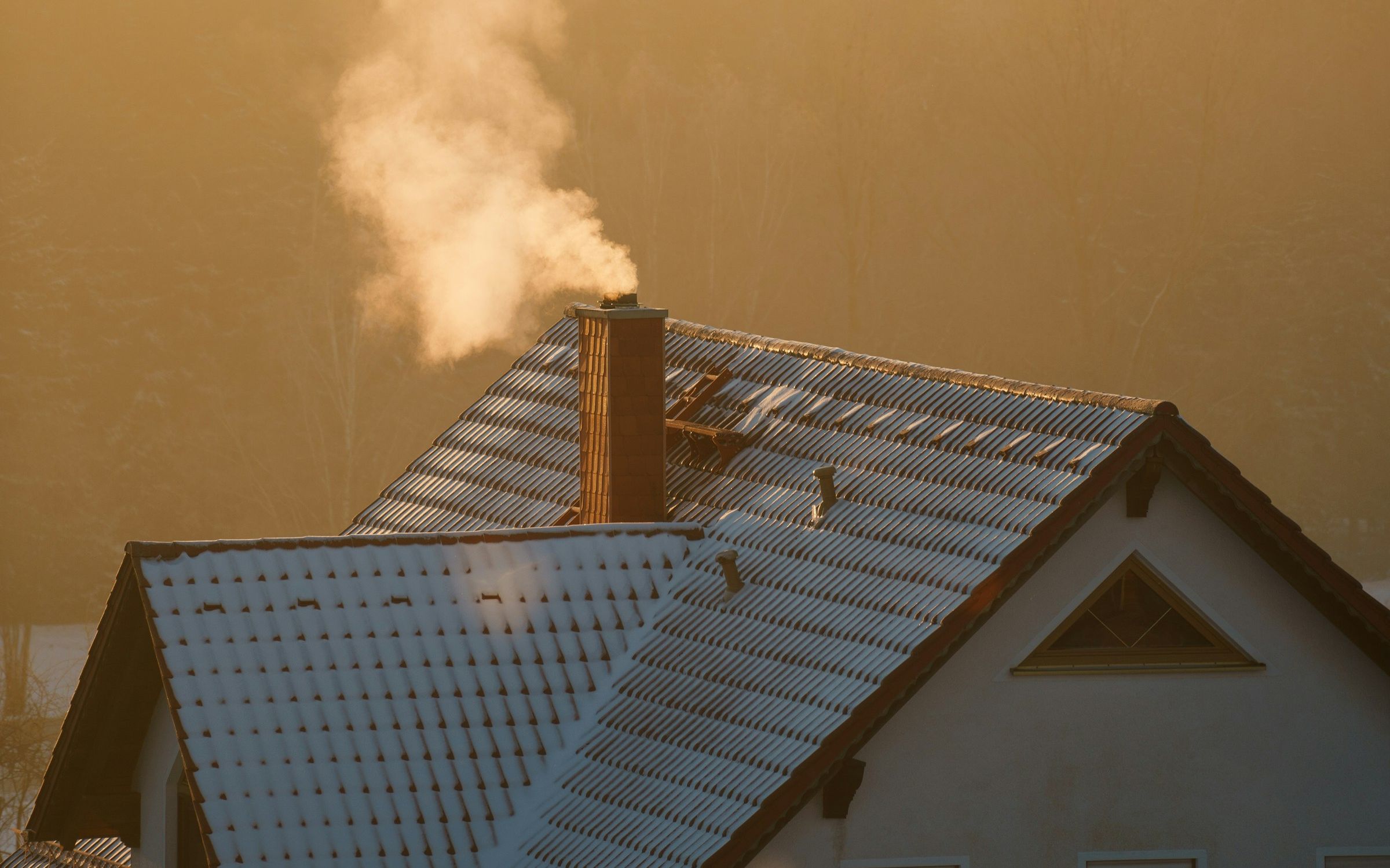 Smoke rising from a chimney over a snowy roof at golden hour